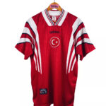Red Adidas football jersey hanging on a wooden hanger, featuring a white crescent and star emblem and bold white stripes on the sleeves with Turkish branding.