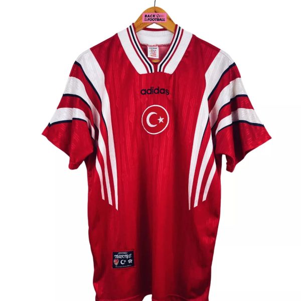 Red Adidas football jersey hanging on a wooden hanger, featuring a white crescent and star emblem and bold white stripes on the sleeves with Turkish branding.
