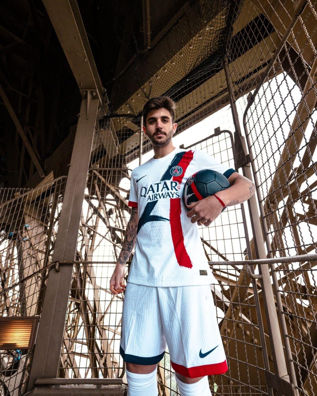 Athlete in a PSG white jersey holding a soccer ball, standing in an industrial metal stairwell. Signage on jersey reads Qatar Airways.