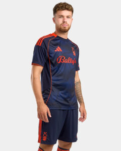 Male model wearing a navy blue football kit with orange trim: short-sleeve jersey showing Bally's sponsor logo and Adidas logo, matching navy shorts with orange stripes.