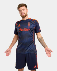 Man modeling a navy soccer jersey and shorts with orange piping and Bally's sponsor text.