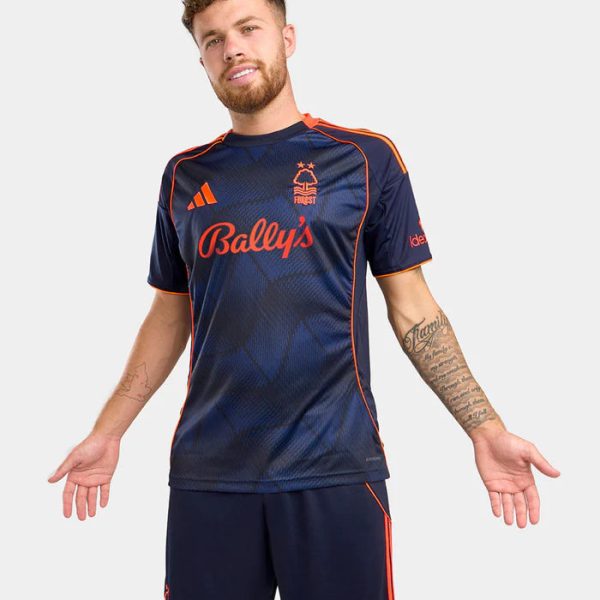 Man modeling a navy soccer jersey and shorts with orange piping and Bally's sponsor text.