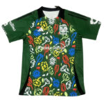 Green soccer jersey with a bold multicolored floral pattern on the front, black side panels, and a black collar; team crest on the left chest and sponsor text across the midsection, with green sleeves.