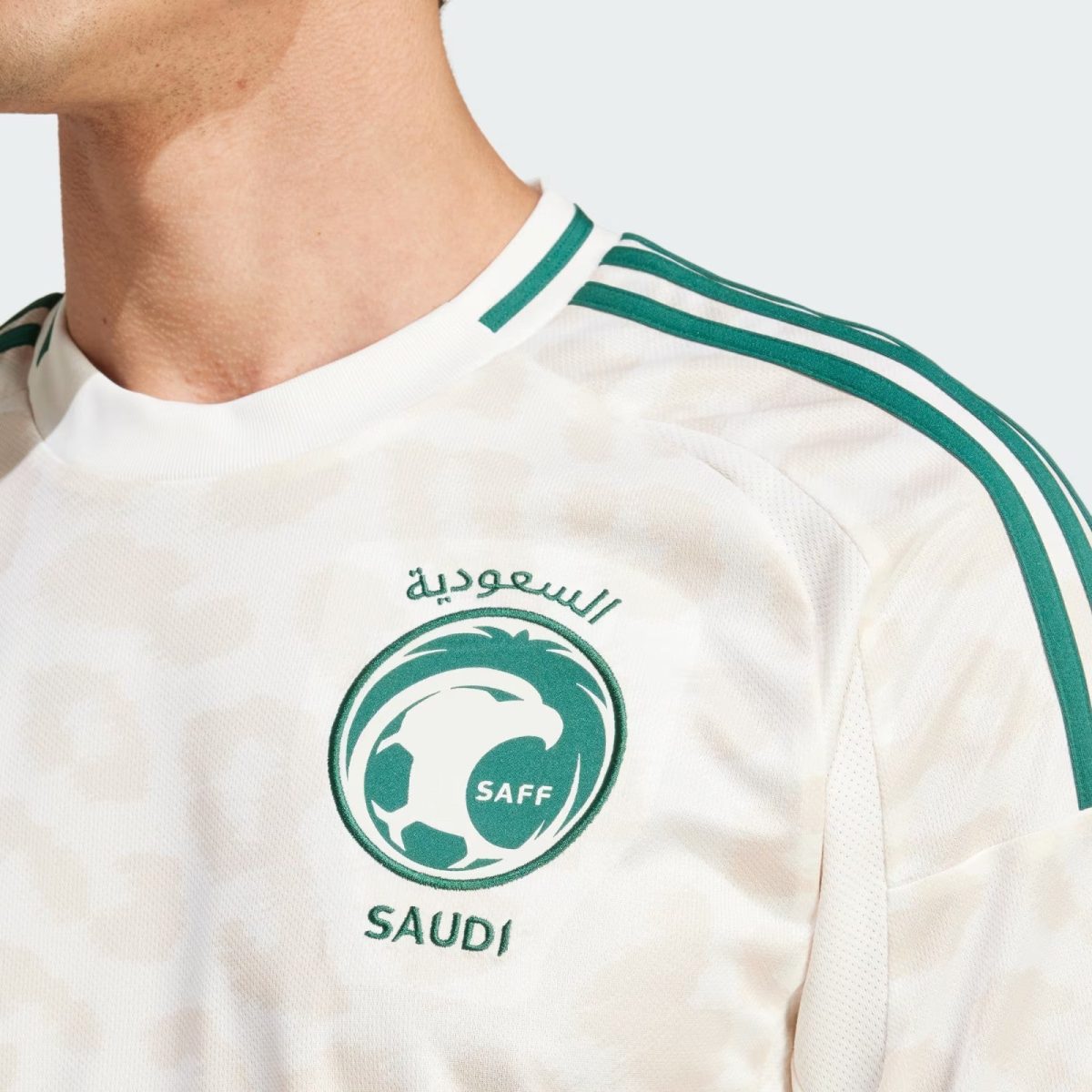 Close-up of a white Saudi football jersey with green stripes along the shoulder and the SAFF logo visible on the chest area, and Arabic script above it