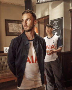Man in a navy jacket and white AIA t-shirt stands in the foreground, looking off to the side, with a second man in a similar jersey in the background inside a cozy room.