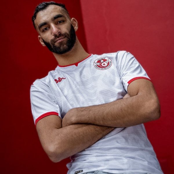 Athlete with a beard in a white jersey with red trim and a circular crest, arms crossed, standing against a red wall.