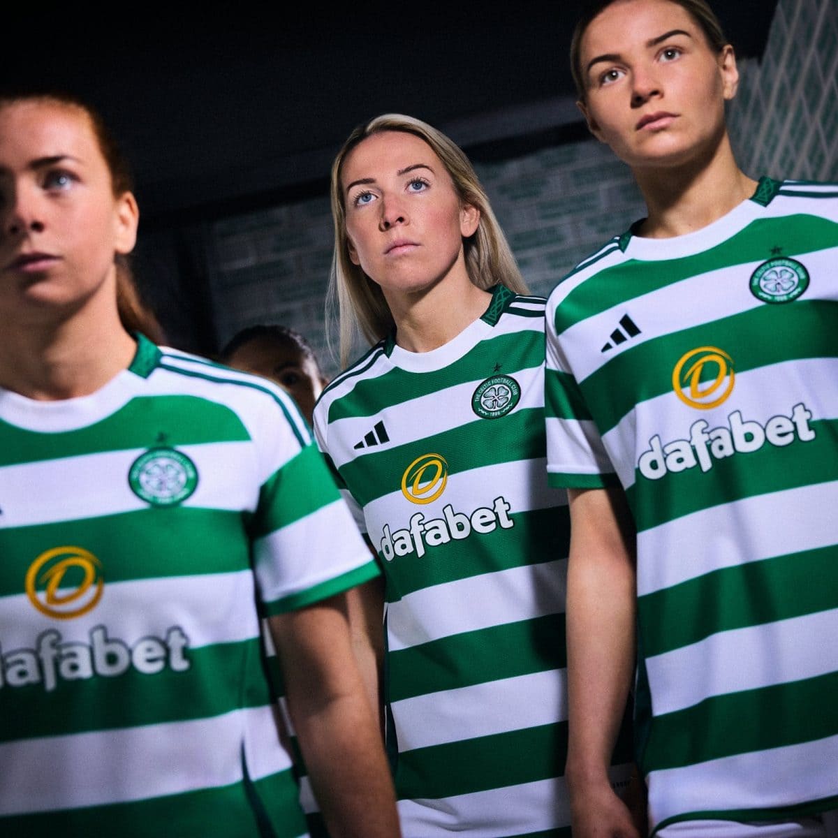 Three women in green and white striped soccer jerseys stand together, looking upward, in a dimly lit setting.