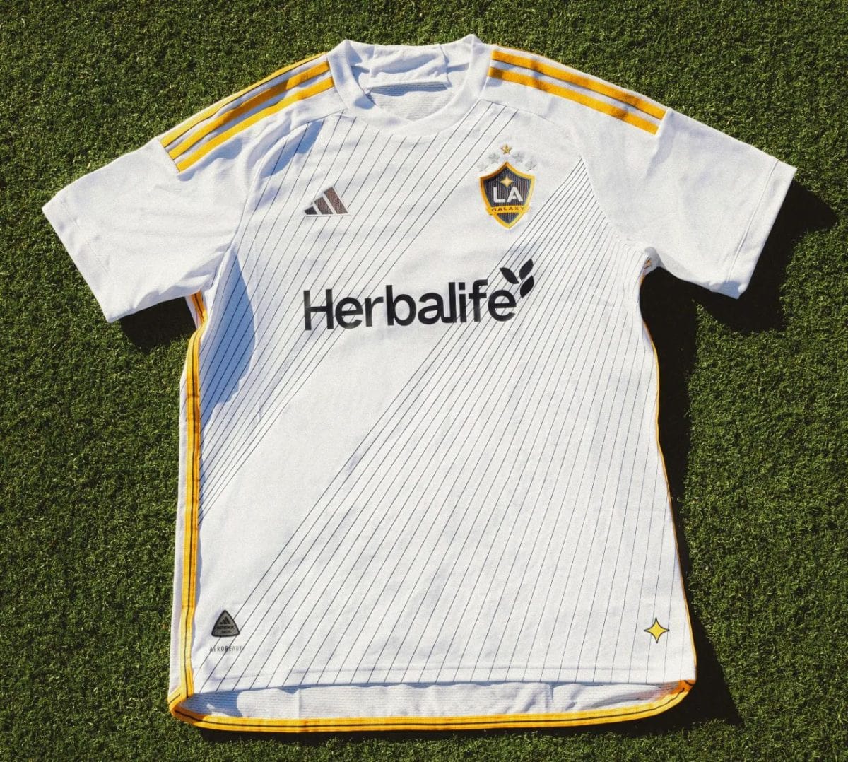 White LA Galaxy soccer jersey on grass with pinstripes, yellow shoulder stripes, and Herbalife sponsor logo front and crest on chest.