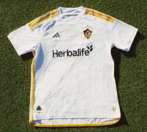UIzqROPN0bxWjFb | UAE SPORT STORE White LA Galaxy soccer jersey on grass with pinstripes, yellow shoulder stripes, and Herbalife sponsor logo front and crest on chest.