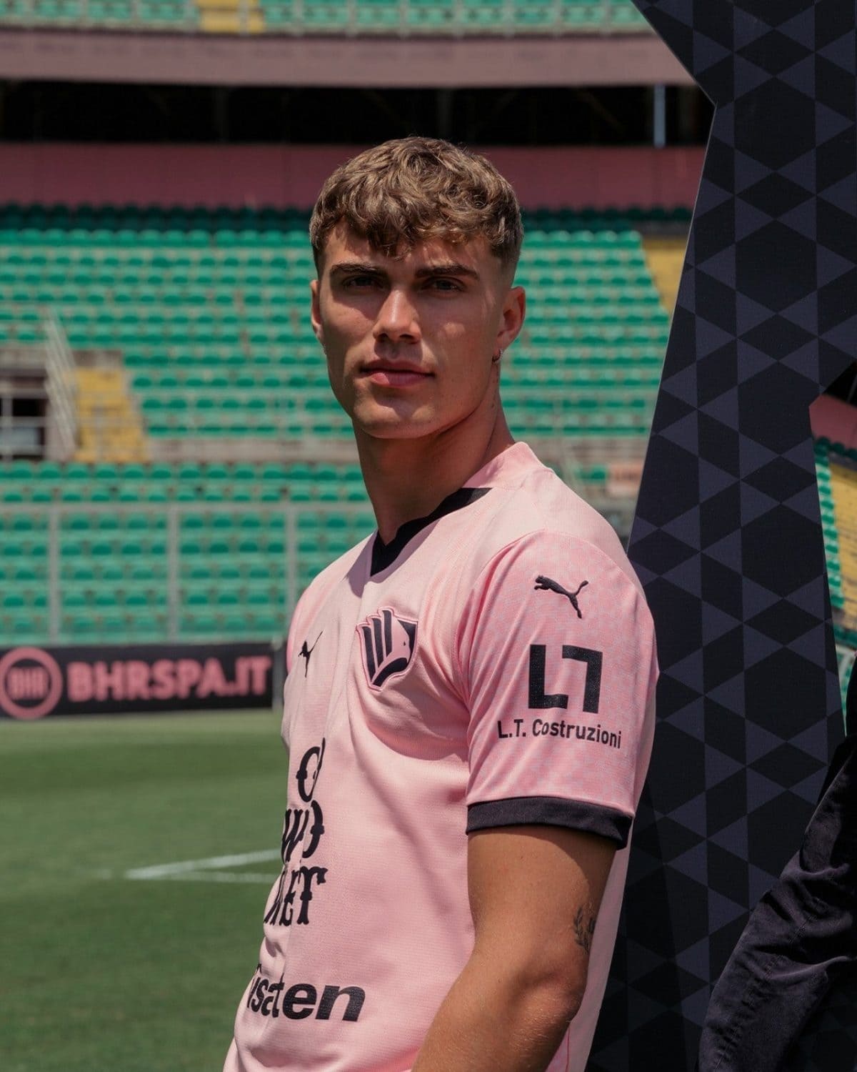 Young man in a pink soccer jersey stands on a green football field with empty green seats in the background.