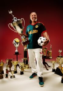UhA15ssbZaw8gxj | UAE SPORT STORE Athlete in a Galaxy jersey smiles among numerous trophies, holding a large trophy and a soccer ball.