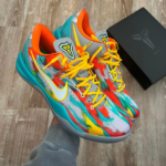 Pair of colorful running shoes with yellow laces and teal accents being held, with a dark box in the background on a wood floor.