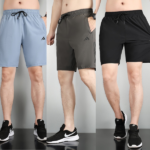 Three male models stand side by side wearing knee-length athletic shorts in light blue, gray, and black with drawstrings and small brand logos on the left thigh; black athletic shoes complete the look.