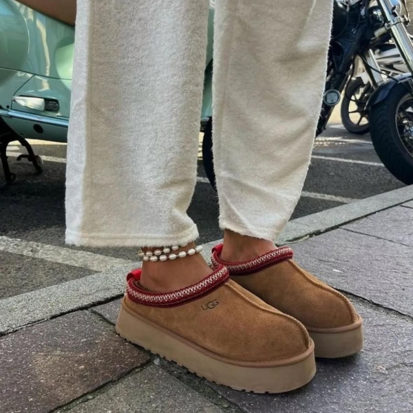 Person's feet in tan UGG-style slippers with red knit trim, white cropped pants, and a pearl anklet, standing on a sidewalk with a car and motorcycle in view.