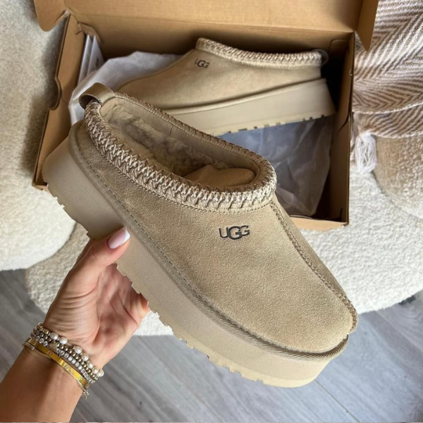 Beige UGG moccasin-style slipper with fuzzy lining held in hand, opened box in background