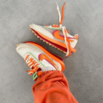 Pair of white sneakers with bright orange accents and green highlights on a gray floor, laces untied.