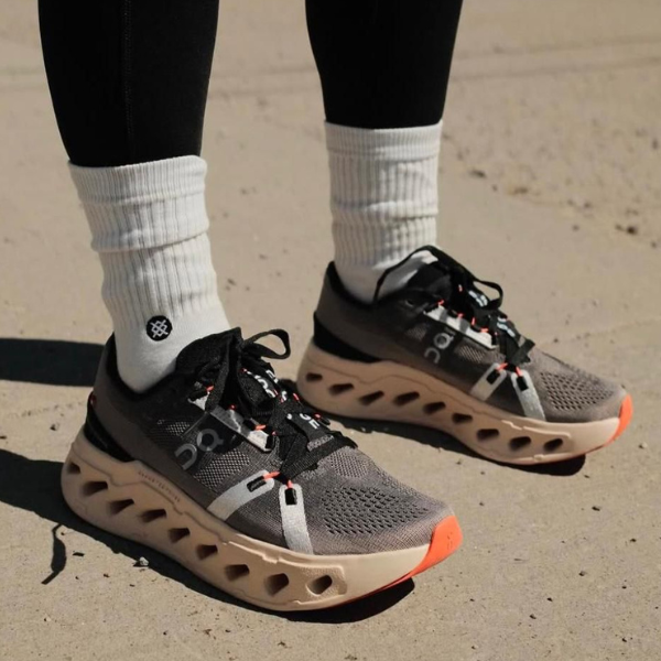 Pair of gray running shoes with a distinctive hollow-cut sole, black and orange laces, worn with white crew socks on pavement.