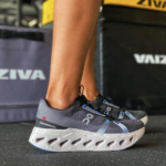Person wearing dark gray On running shoes with white cloud-like sole standing on a gym floor.