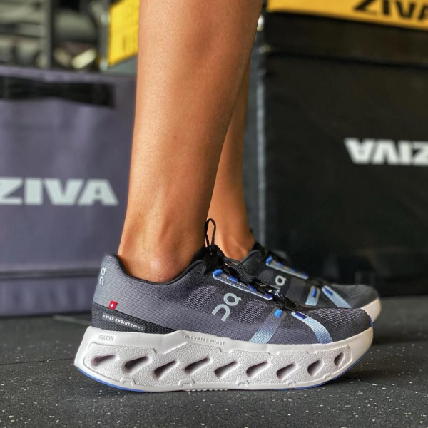 Person wearing dark gray On running shoes with white cloud-like sole standing on a gym floor.
