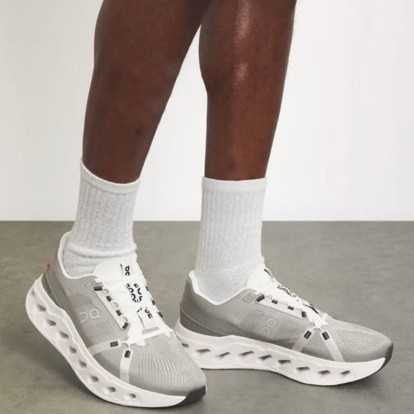 Gray running shoes with white sole and sculpted cutouts, worn with white crew socks on a person’s feet, indoor setting.