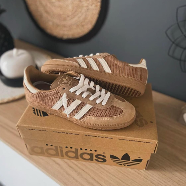 Brown Adidas sneakers with white stripes resting on a cardboard Adidas box on a wooden surface indoors.