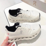 Pair of white running shoes with black inner lining displayed on a white surface next to a gray box, featuring the On logo on the sides and midsole holes for cushioning.