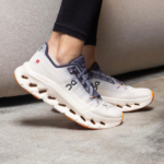 Pair of gray and white running shoes with orange accents worn by someone standing on a carpeted floor.