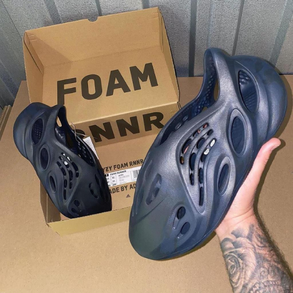Tattooed hand holding a gray Foam Runner shoe in front of a cardboard box labeled FOAM RUNNER(s).