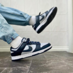 Person wearing light-wash jeans and white socks kicks a navy and white sneaker mid-air on a tiled floor, showing the side of the shoe.