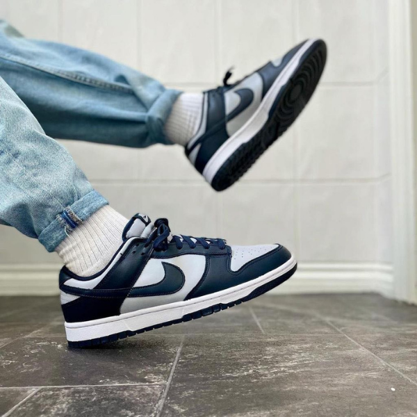 Person wearing light-wash jeans and white socks kicks a navy and white sneaker mid-air on a tiled floor, showing the side of the shoe.