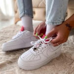 Person tying white Nike sneakers with neon green nails, a jeweled lace charm visible on the laces, indoors on a beige rug.
