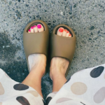 Top-down view of feet wearing olive-green slide sandals on a concrete surface, showing pink and orange pedicure nails.
