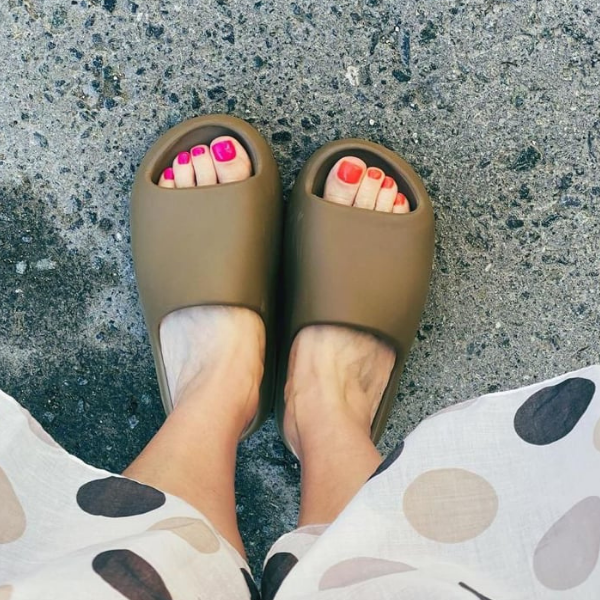 Top-down view of feet wearing olive-green slide sandals on a concrete surface, showing pink and orange pedicure nails.