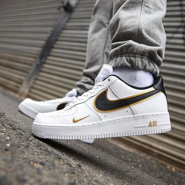 White Nike Air Force 1 sneakers with black and gold swoosh, worn with gray joggers, standing on a curb.