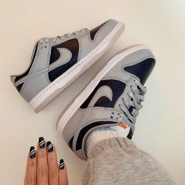 Gray and navy Nike sneakers with white soles resting on a light surface; a hand with black-and-white striped nails sits nearby.