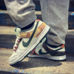 Close-up of white and gray Nike sneakers with camouflage panels and a green swoosh on concrete stairs with gray joggers nearby.