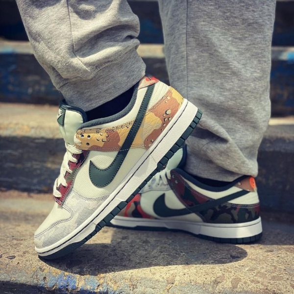 Close-up of white and gray Nike sneakers with camouflage panels and a green swoosh on concrete stairs with gray joggers nearby.