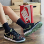 Person wearing black camouflage Nike sneakers with a green swoosh, seated on a wooden floor with cardboard shoe boxes in the background.