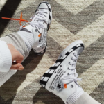 Two white sneakers with black stripes on a beige rug, laces tied, orange tag on the left shoe with text visible on the sides of both sneakers.