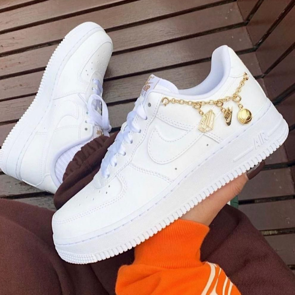 White Nike Air Force 1 sneakers with a gold charm bracelet attached, shown on a wooden deck.