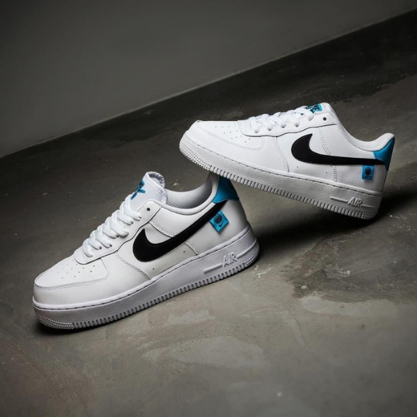 White Nike Air sneakers with a black swoosh and blue accents on a gray floor, photographed from a low angle.