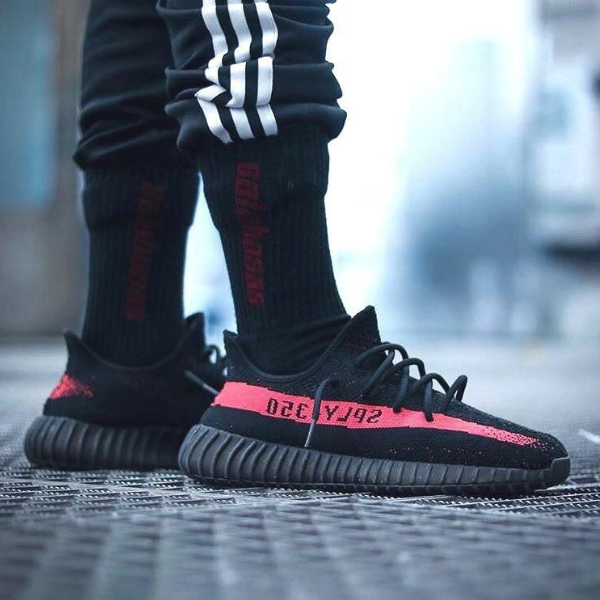 Close-up of a person wearing black and red sneakers with a pink stripe on a metal grate, urban setting in the background.
