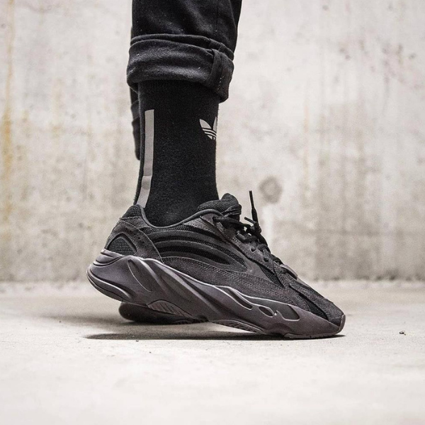 Black chunky sneaker on a foot wearing a black sock with a white logo, standing on a concrete floor.