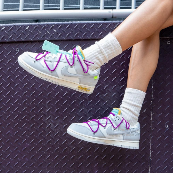 Person wearing light gray sneakers with bright pink laces and white socks, legs resting on a purple diamond-plate surface with tags on the shoes nearby.