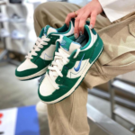 Pair of green and white Nike sneakers held by a person wearing khaki pants, blue swoosh visible in the foreground.