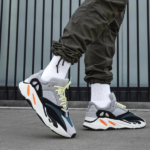 Pair of gray and black chunky sneakers with lime laces, white socks, and olive joggers on a concrete surface.