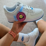 White Nike Air Force 1 sneakers with light blue accents, held by a person wearing a pink smartwatch.