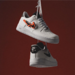 Two white Nike Air sneakers hanging by their laces against a dark red backdrop, with an orange tag on the top shoe.