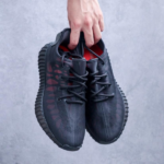 A person holds two black lace-up sneakers by the top, showing the side and red inner lining against a gray background.