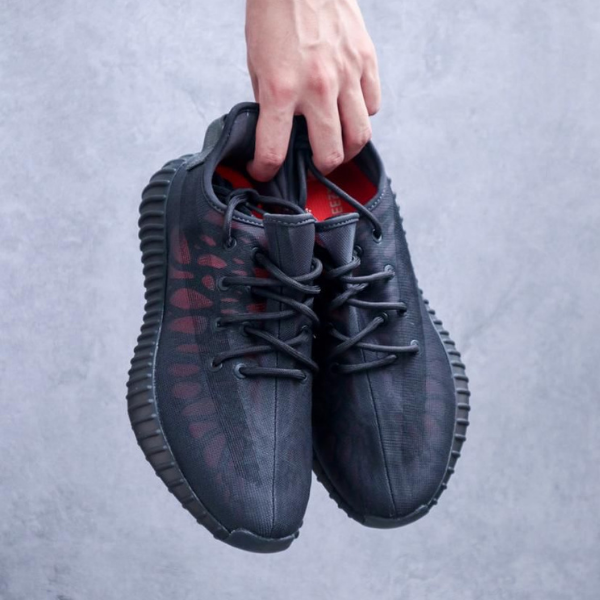A person holds two black lace-up sneakers by the top, showing the side and red inner lining against a gray background.